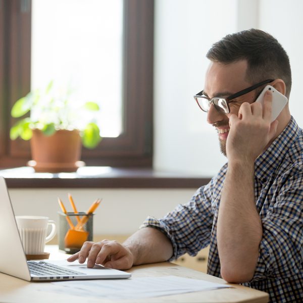 Smiling male worker talking over cell, consulting client online, typing message on laptop, writing email, giving help, assistance. Freelancer person speaking on phone, working. Concept of multitasking